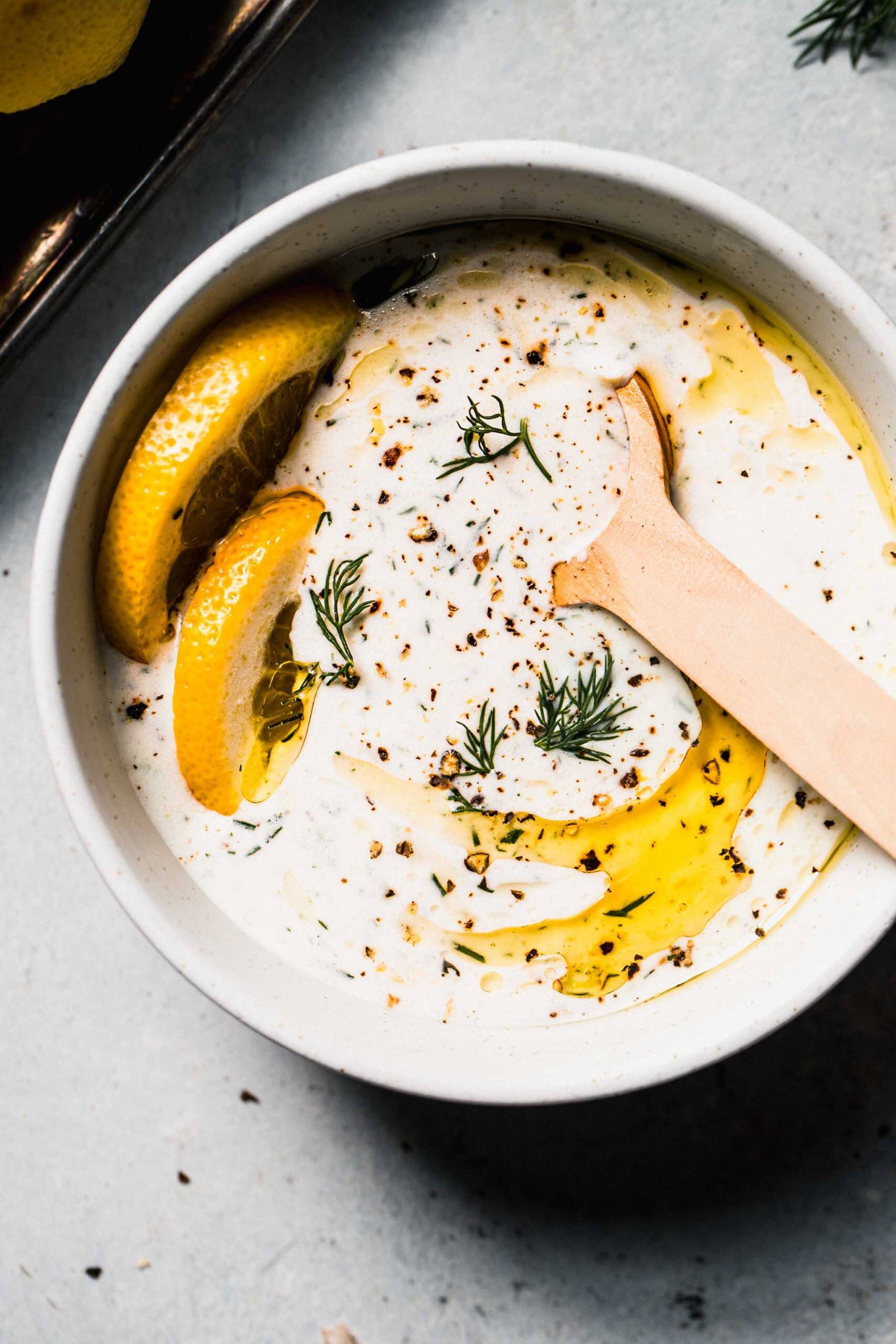 Bowl of yogurt dill sauce with spoon and slices of lemon, drizzled with olive oil.