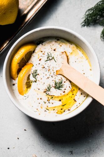 Bowl of yogurt dill sauce with spoon and slices of lemon, drizzled with olive oil.