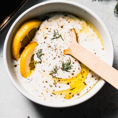 Bowl of yogurt dill sauce with spoon and slices of lemon, drizzled with olive oil.