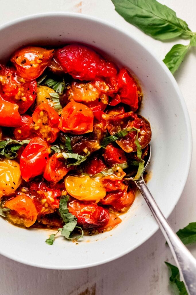 Air fried tomatoes in white bowl with serving spoon topped with basil sprigs.
