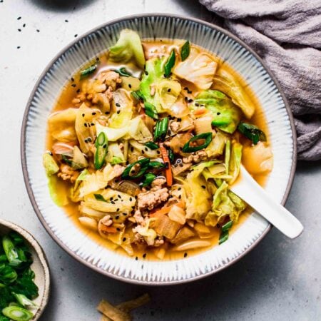 Bowl of egg roll soup on counter next to small bowls of sesame seeds, green onions and wonton strips.