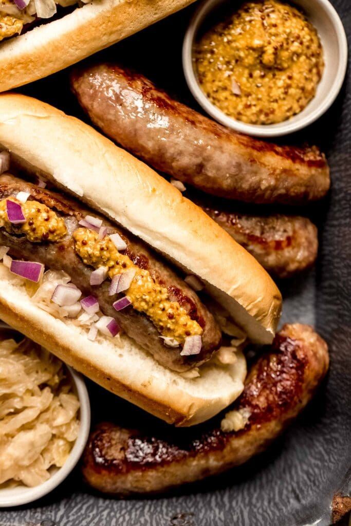 Bratwurst in buns arranged on serving tray next to small bowls of mustard and sauerkraut.