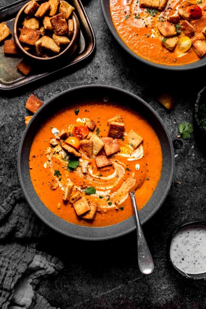 Two bowls of indian tomato soup on counter next to bowl of naan croutons and creamy yogurt sauce. 