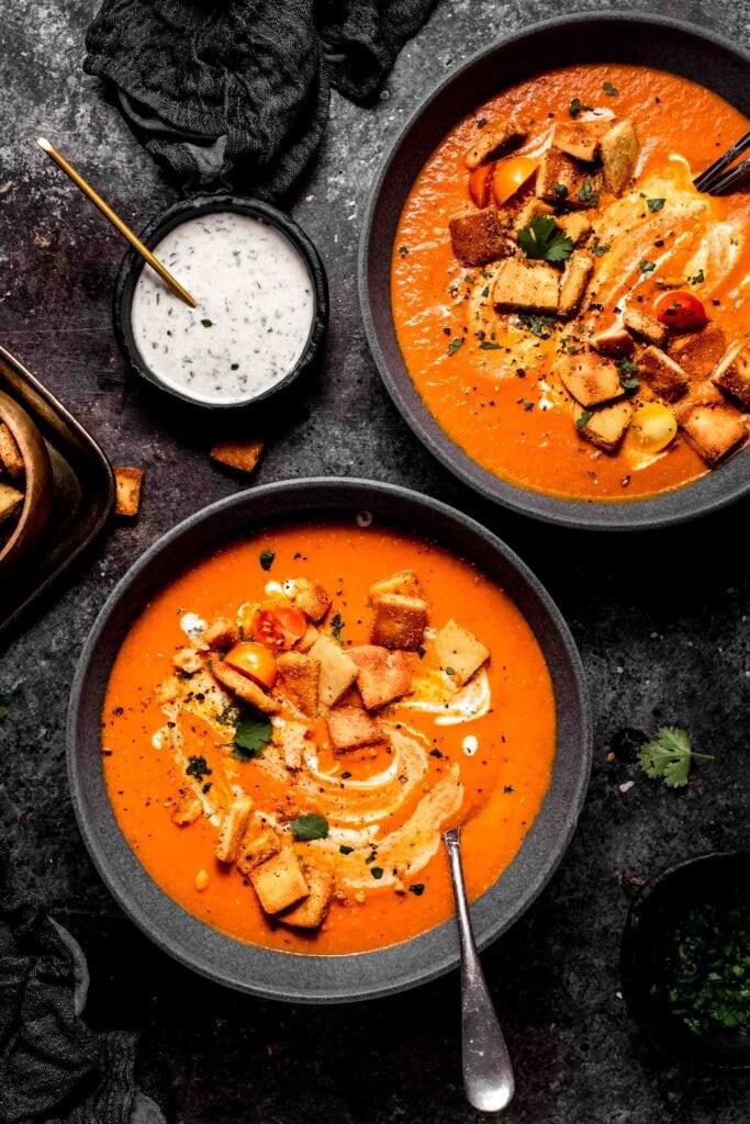Two bowls of indian tomato soup on counter next to bowl of naan croutons and creamy yogurt sauce.