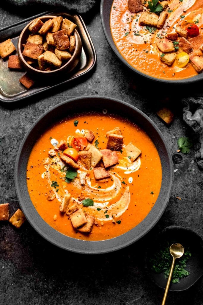 Two bowls of indian tomato soup on counter next to bowl of naan croutons and creamy yogurt sauce. 