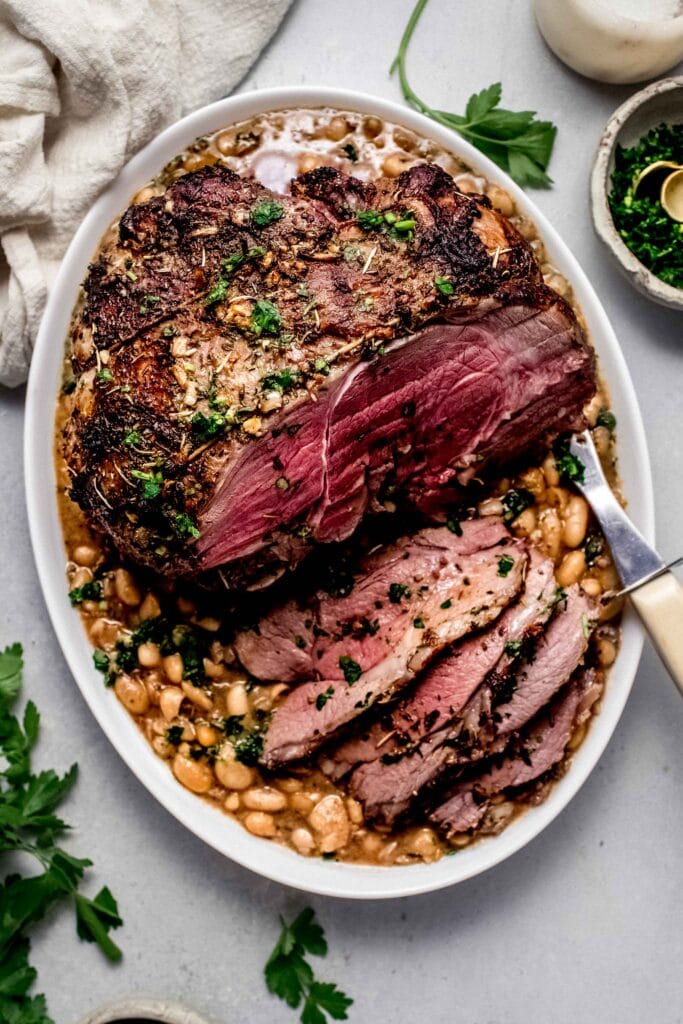 Prepared leg of lamb in serving dish with white beans underneath. 