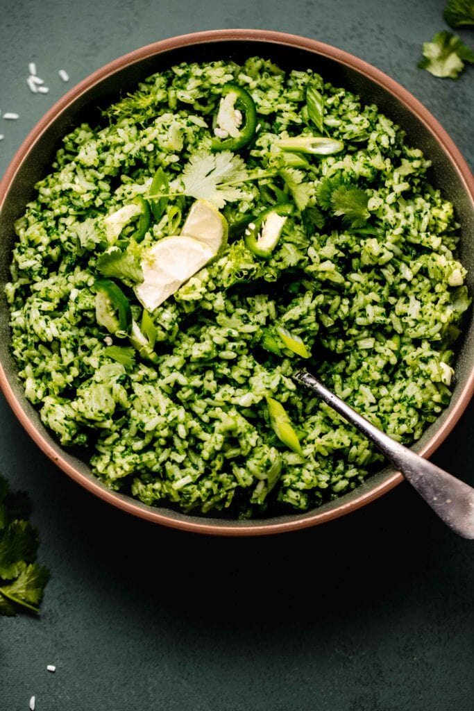 Green rice in green bowl with spoon topped with lime wedges and cilantro leaves.