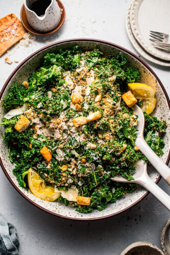 Overhead shot of kale salad tossed in bowl and topped with croutons and lemon wedges.