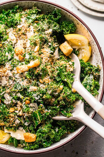 Overhead close up of simple kale salad in bowl with salad serving utensils.