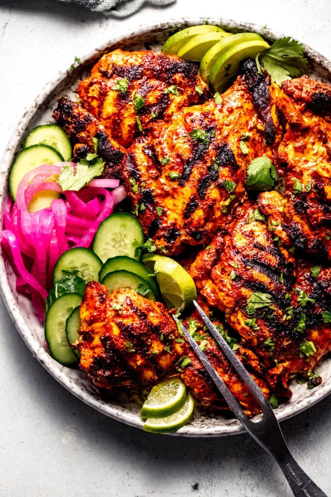 Grilled achiote chicken arranged on serving plate with pickled onions, cucumbers and lime wedges.