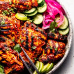 Grilled achiote chicken arranged on serving plate with pickled onions, cucumbers and lime wedges.