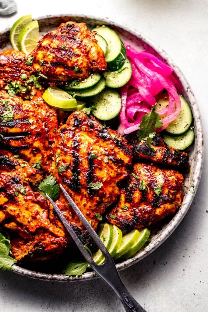 Grilled achiote chicken arranged on serving plate with pickled onions, cucumbers and lime wedges.