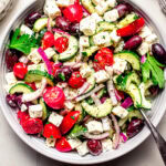 Overhead shot of greek tomato cucumber salad in white bowl next to bowl of feta.
