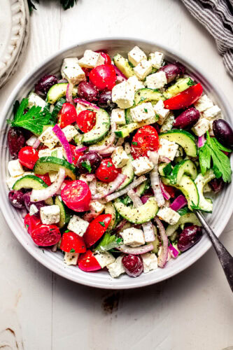 Overhead shot of greek tomato cucumber salad in white bowl next to bowl of feta.