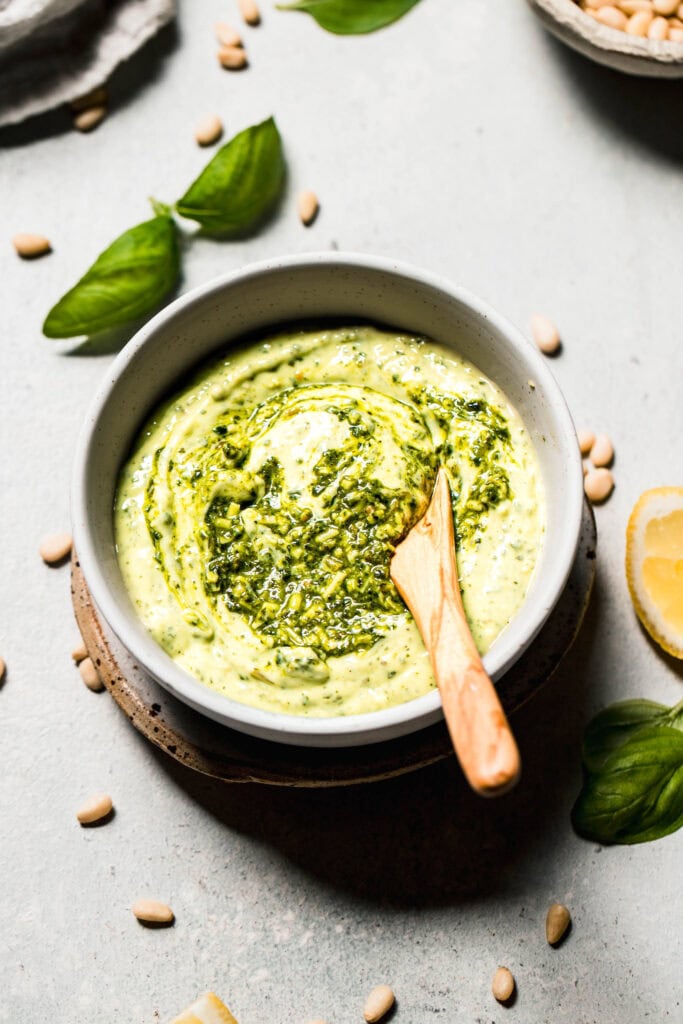 Side view of pesto mayo in small white bowl with wooden spoon.