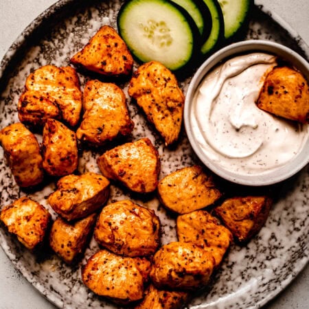 Air fryer chicken bites arranged on plate with bowl of dipping sauce and cucumber slices.
