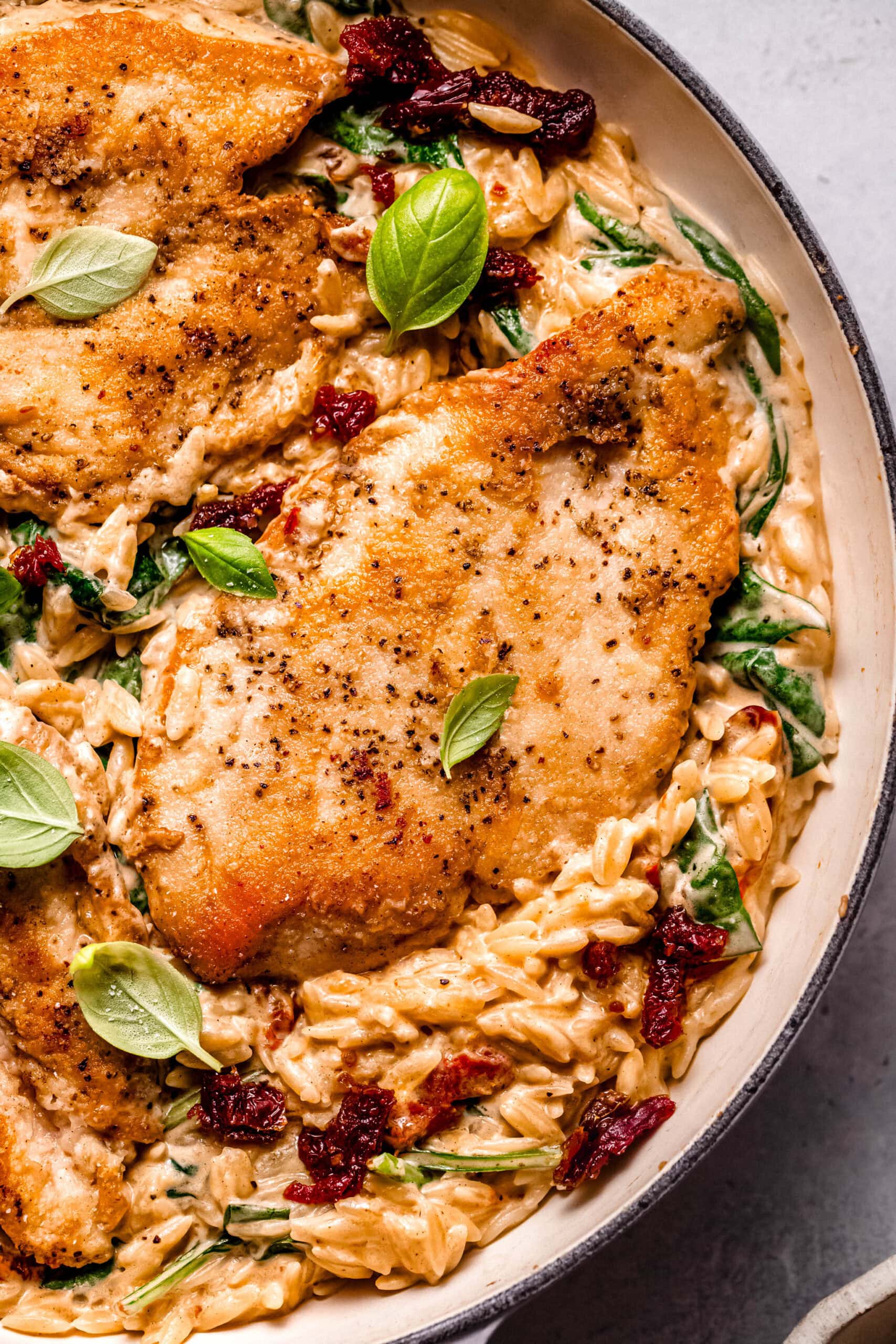 Overhead close up of prepared sun dried tomato orzo topped with chicken breasts in skillet.