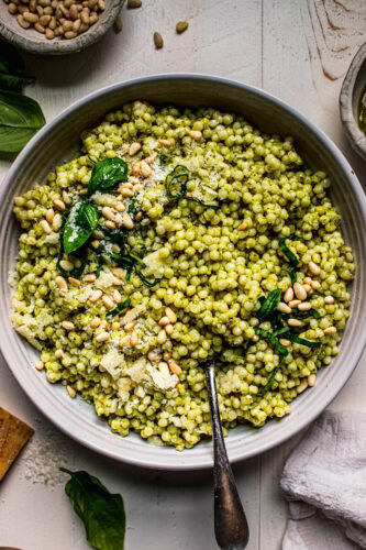 Prepared pesto parmesan couscous in serving bowl topped with basil and pine nuts.