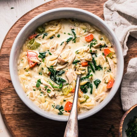Overhead shot of creamy orzo chicken soup in white bowl with spoon on wooden cutting board backdrop.
