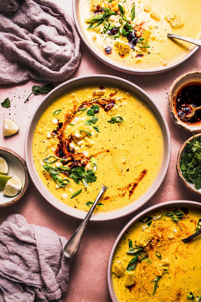 THREE BOWLS OF CREAMY CURRY CORN CHOWDER ON PINK TABLE NEXT TO BOWLS OF CHILI CRISP AND GREEN ONIONS. 