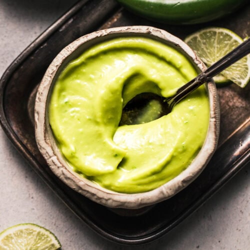 Avocado hot sauce in small bowl with spoon next to lime wedges.