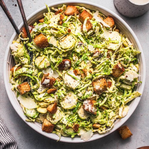 Overhead shot of brussel sprout caesar salad in white serving bowl with serving spoons.
