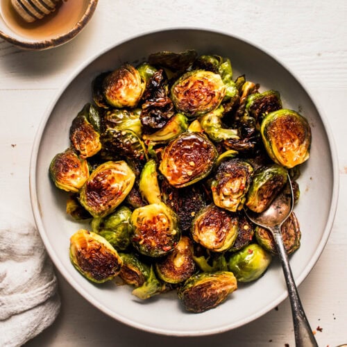 Overhead shot of honey balsamic brussels sprouts in white bowl next to bowl of honey and bowl of balsamic glaze.
