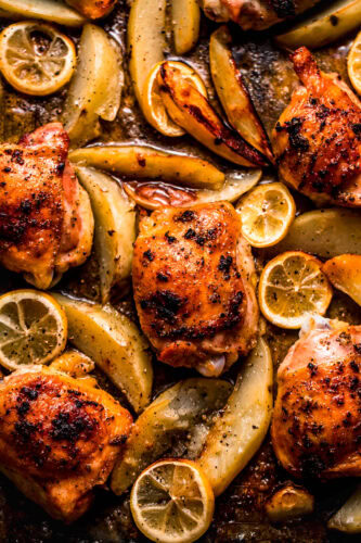Close up of baked chicken and potatoes on baking sheet scattered with lemon slices.