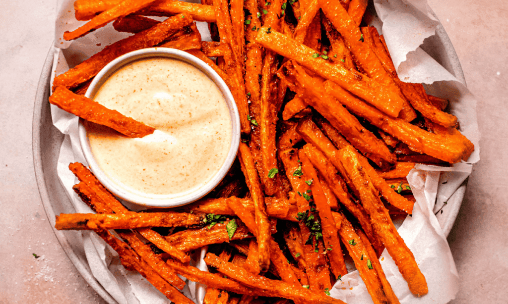 Overhead close up horizontal image of Rustic basket of oven-baked carrot fries with parchment, reaching into a creamy sriracha mayo bowl.