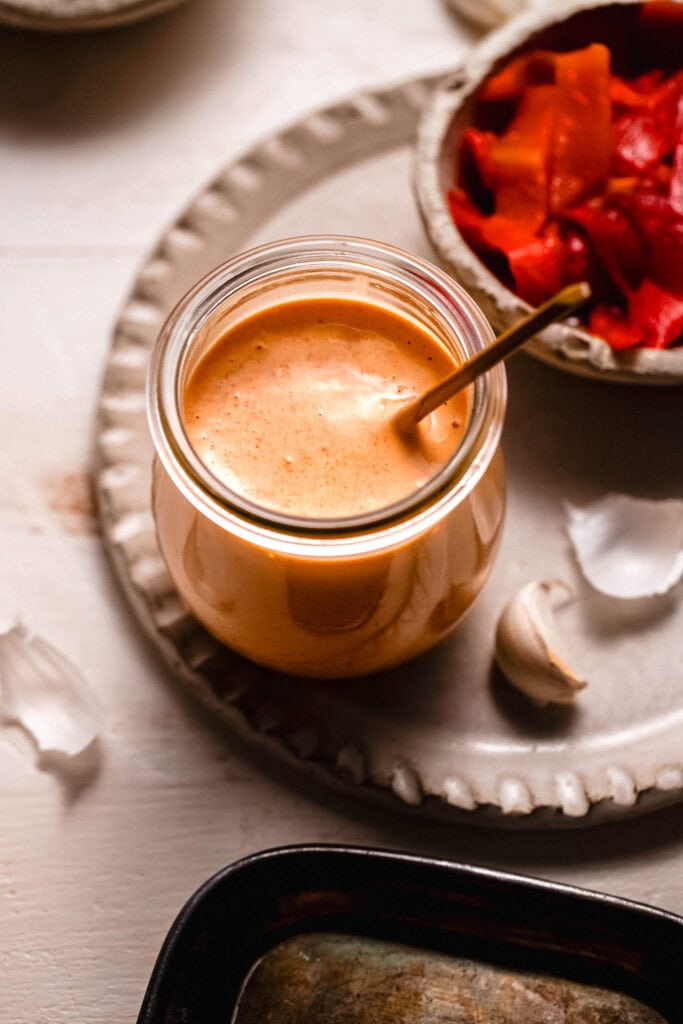Red pepper aioli in small bowl with gold spoon next to bowl of roasted red peppers.