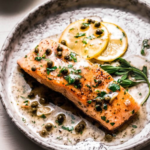 Side view of salmon piccata on plate with lemon slices and sprig of parsley.