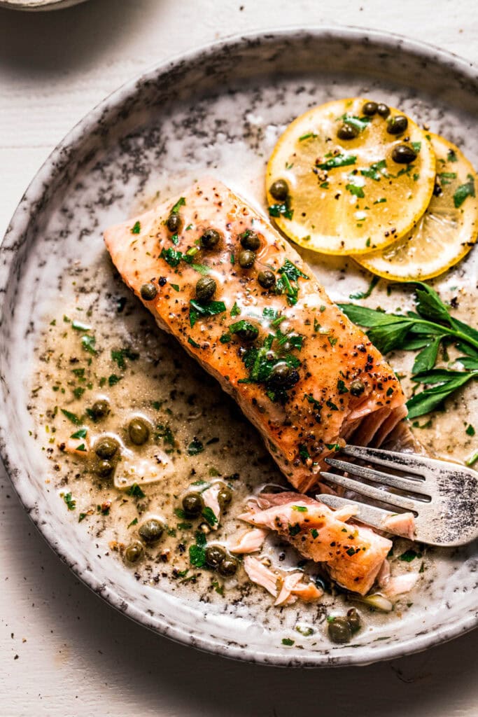 Fork slicing into salmon piccata on plate.