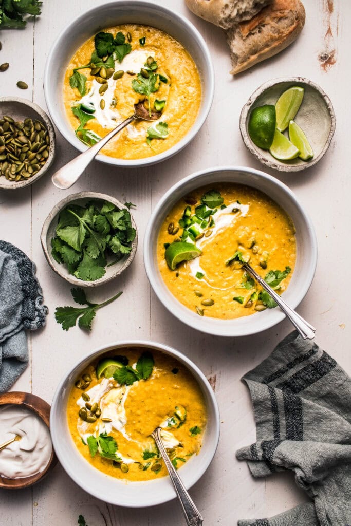 Three bowls of creamy poblano soup on counter next to bowls of cilantro, lime wedges, and pumpkin seeds.