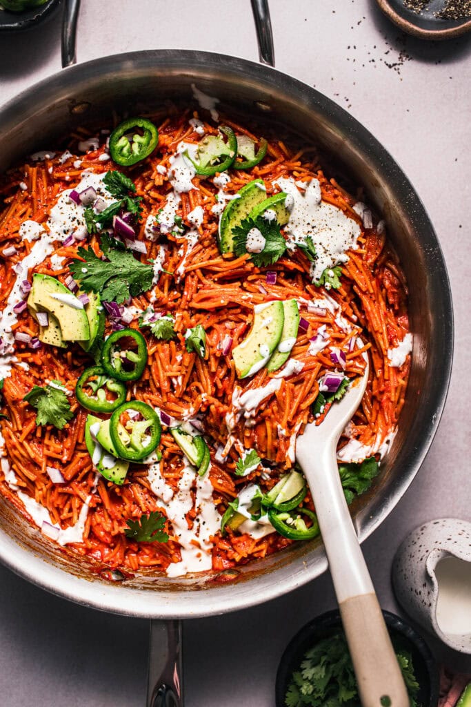 Close up of finished fideo seco in skillet topped with sour cream, avocados, red onion and jalapeno.