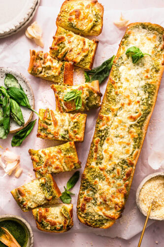 Pesto garlic bread loaves on counter being sliced into topped with basil leaves.