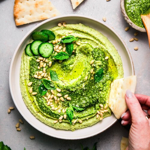Overhead shot of piece of naan dipping into bowl of pesto hummus.