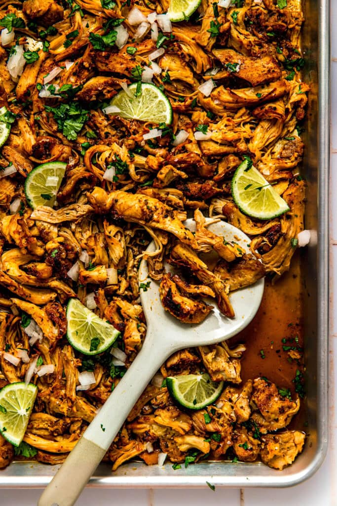 Shredded chicken carnitas on baking sheet with lime wedges and cilantro.