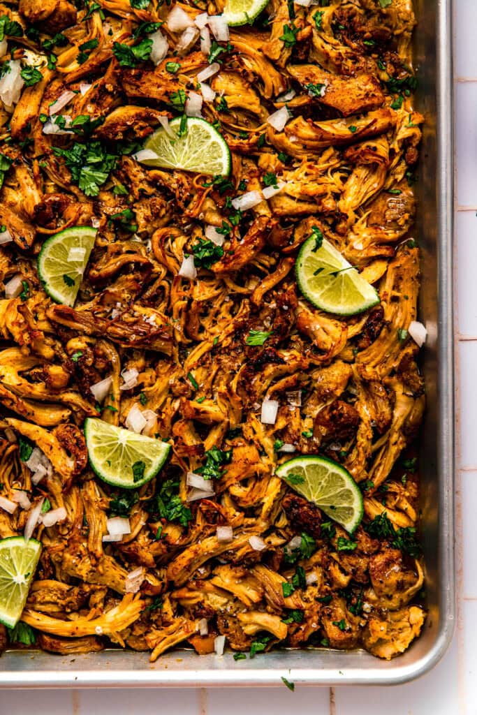 Shredded chicken carnitas on baking sheet with lime wedges and cilantro.