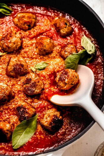 Close up of finished chicken ricotta meatballs in skillet with serving spoon. Bathed in tomato sauce, parmesan and basil leaves.