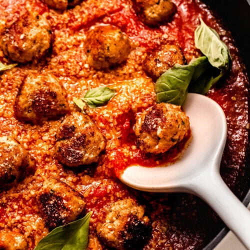 Close up of finished chicken ricotta meatballs in skillet with serving spoon. Bathed in tomato sauce, parmesan and basil leaves.