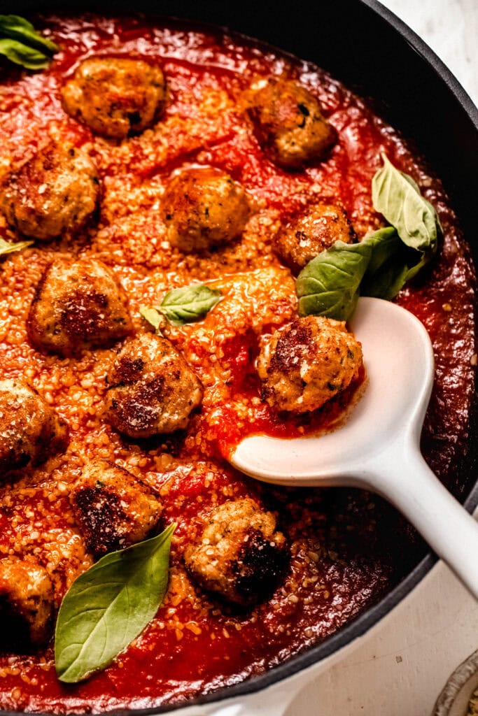 Close up of finished chicken ricotta meatballs in skillet with serving spoon. Bathed in tomato sauce, parmesan and basil leaves.