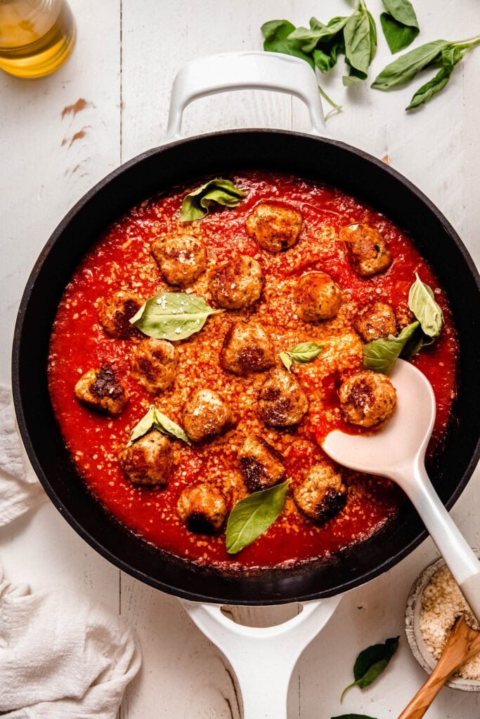 Finished chicken ricotta meatballs in skillet with serving spoon. Bathed in tomato sauce, parmesan and basil leaves.