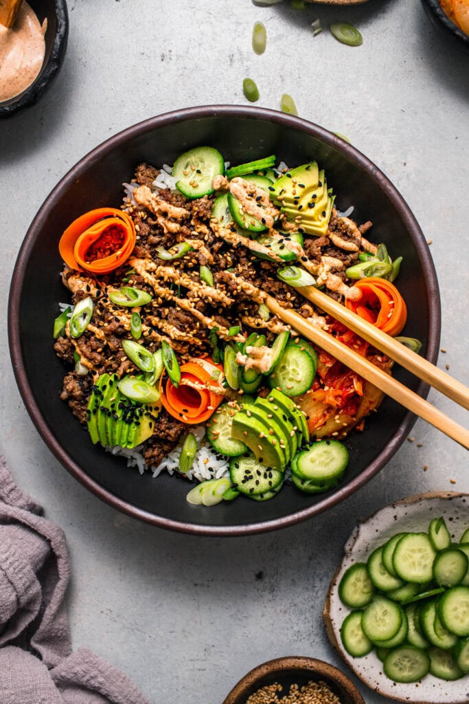 Assembled korean beef bowl on counter next to small bowl of sliced cucumber.