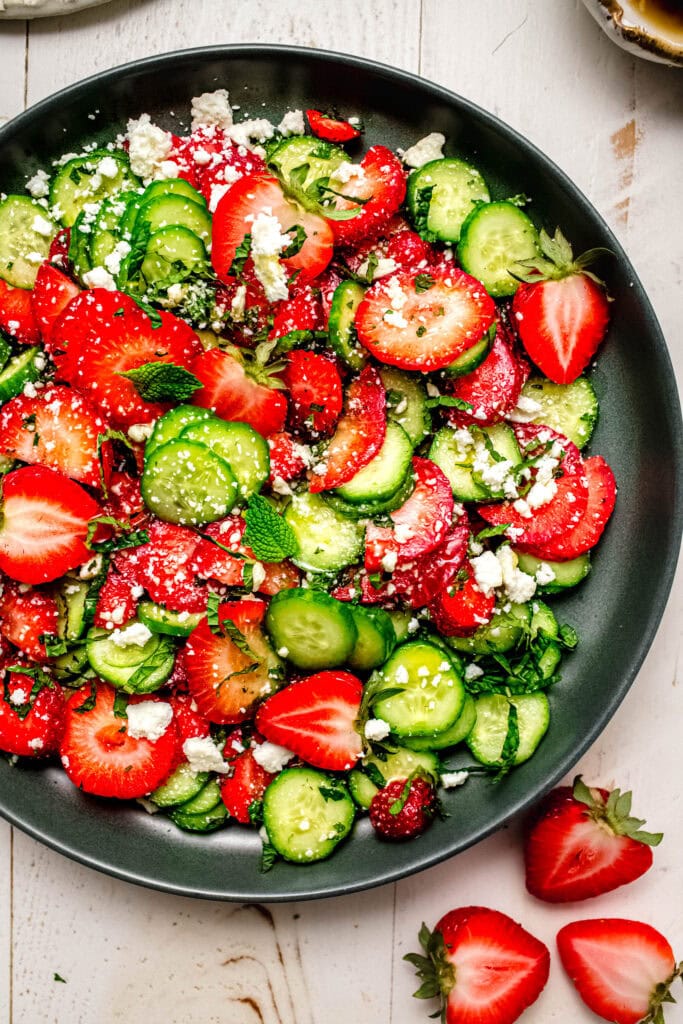 Overhead close up of Fresh cucumber and strawberry salad on a green serving platter, surrounded by sliced fruit and vegetables.