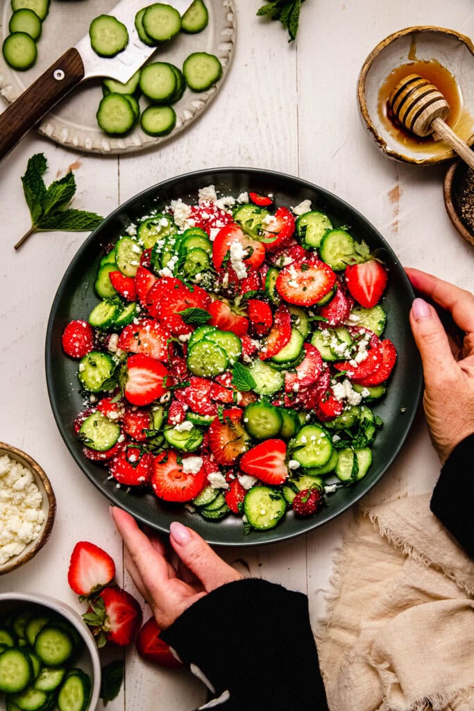 Hands holding Strawberry cucumber salad on green serving plate next to sliced cucumbers and strawberries.