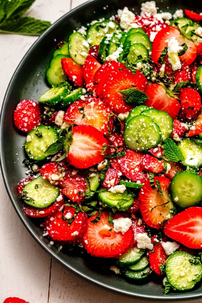 Overhead close up of Fresh cucumber and strawberry salad on a green serving platter, surrounded by sliced fruit and vegetables.