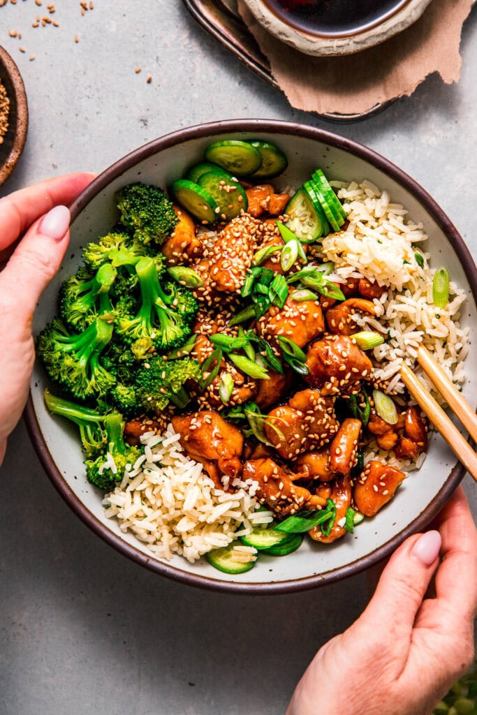 Hands holding teriyaki chicken rice bowl with chopsticks.
