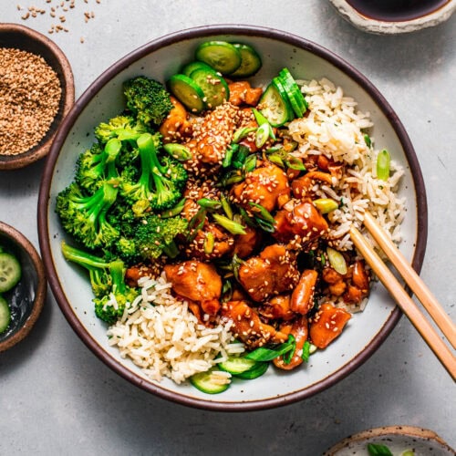 Asian-inspired teriyaki chicken rice bowl served with cucumber slices, chopped green onions, and sesame seed garnish.