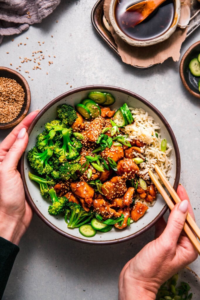 Hands holding teriyaki chicken rice bowl with chopsticks.
