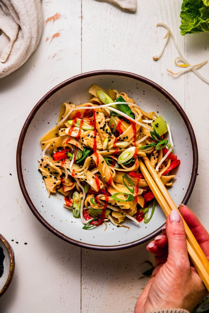 Hands dipping chopsticks into bowl of chicken chow fun.